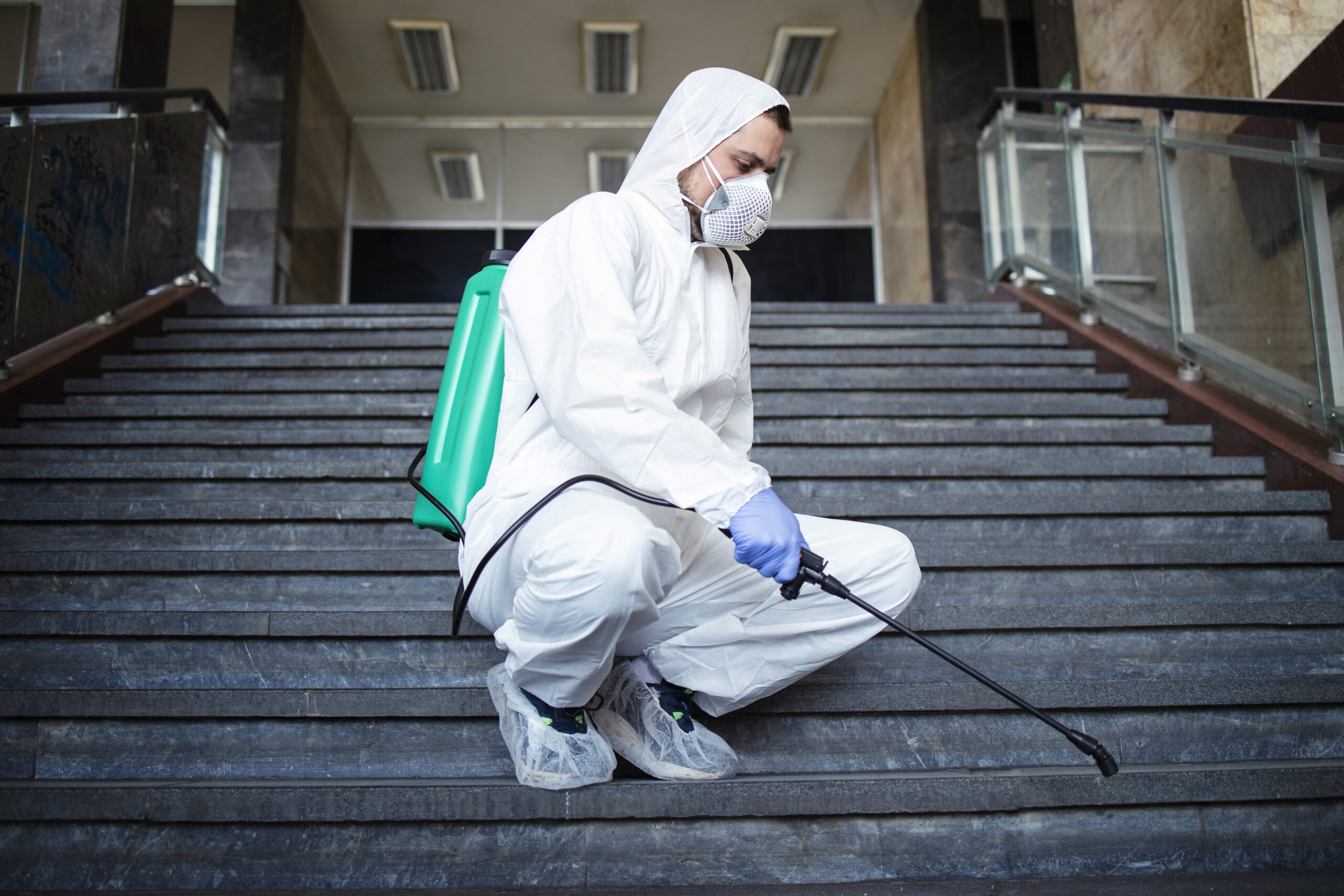 Person in white chemical protection suit disinfecting public corridors and steps to stop spreading highly contagious corona virus.